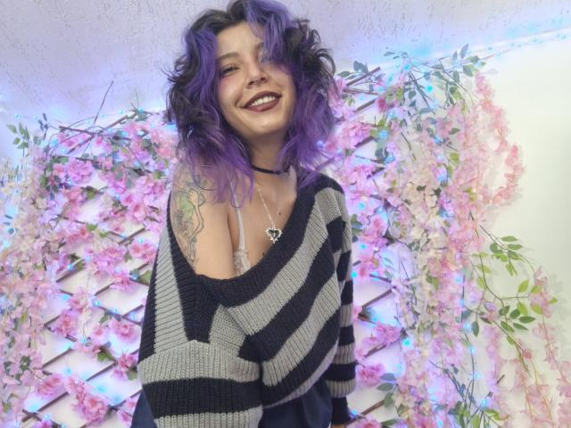 DarckVelvet with purple hair, smiling, wearing a black and gray striped sweater, showing a tattoo on her right shoulder, and a heart necklace, standing in front of pink flowers.
