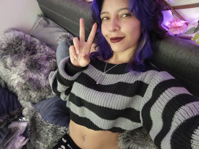DarckVelvet, avec des cheveux violets, un pull rayé noir et gris, affichant un signe de paix, du rouge à lèvres foncé, un anneau au nez et un piercing au nombril, assise sur un canapé gris..
