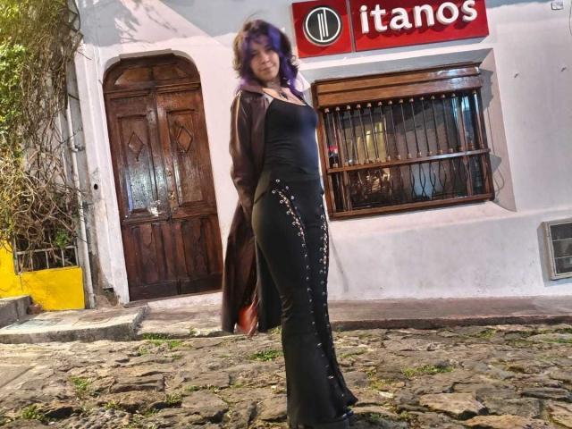 DarckVelvet si trova in una strada acciottolata, indossa un abito nero aderente con paillettes argentate, un cardigan marrone, e i suoi capelli viola sono leggermente ondulati. Sullo sfondo si vede una porta di legno..