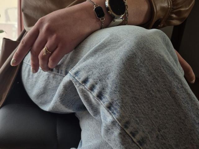 LoriEla's hand with a gold ring and black watch rests on her blue jeans, showing a brown leather jacket. Her wrist is slightly bent, fingers relaxed.