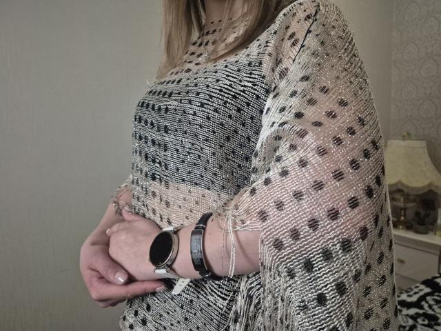 LoriEla's hands are crossed in front of her, wearing a fringed, black-and-white patterned knit top, with a black wristwatch on her left wrist.