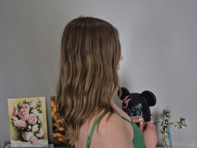 LoriEla with wavy brown hair, wearing a green top, holding a black Minnie Mouse head, her back to the camera, and a flower painting on a shelf.