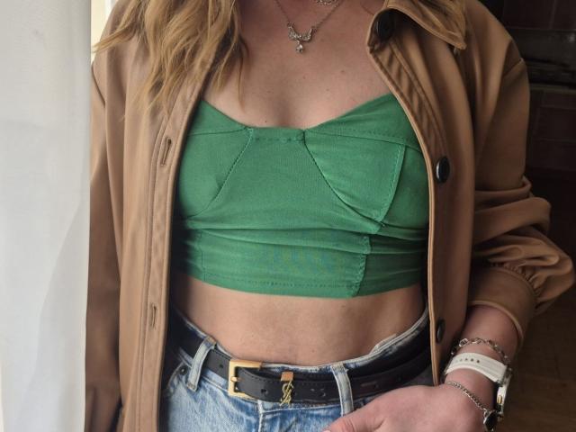 LoriEla with green crop top showing her tits, tan jacket, blue jeans, black belt, and jewelry, including necklace and watch, with blonde hair.