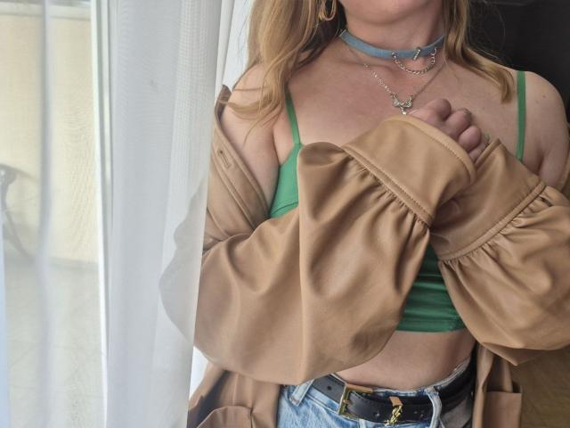 LoriEla with green bra, tan jacket, blue choker, and denim jeans, holding jacket off shoulders, showing tan belt with gold buckle, blonde hair, and gold hoop earrings.