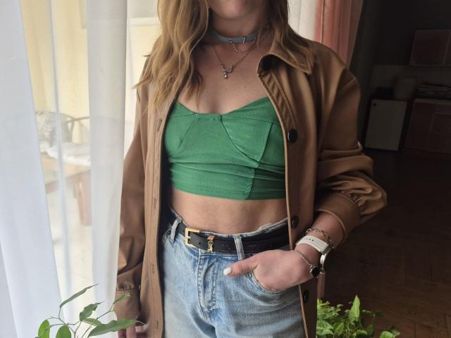 LoriEla with wavy brown hair, green crop top showing small tits, tan jacket, blue jeans, black belt, and chokers, standing by a window with sunlight.