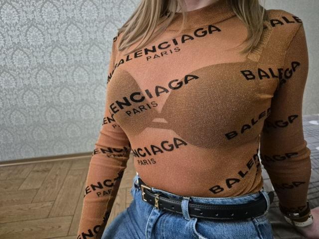 LoriEla's orange Balenciaga top shows her big tits through a brown, heart-shaped cutout. She's wearing high-waisted blue jeans with a black belt.