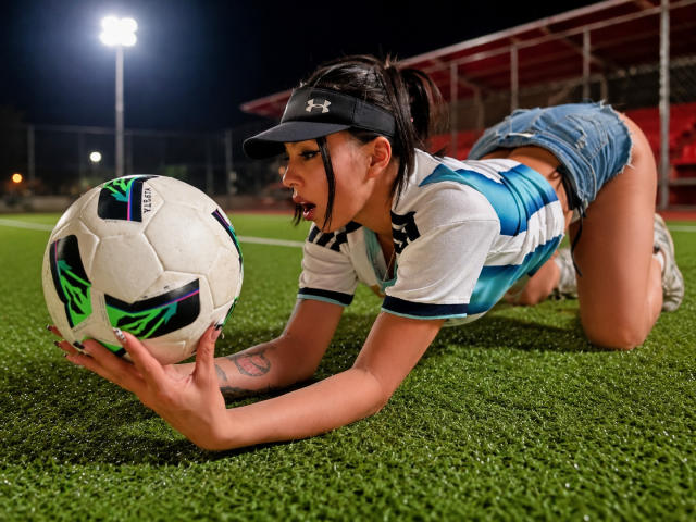 NenaBlack kniet, trägt ein weiß-blaues Fußballtrikot, Jeansshorts und eine schwarze Kappe und hält einen Fußball mit grün-schwarzen Mustern auf grünem Rasen..