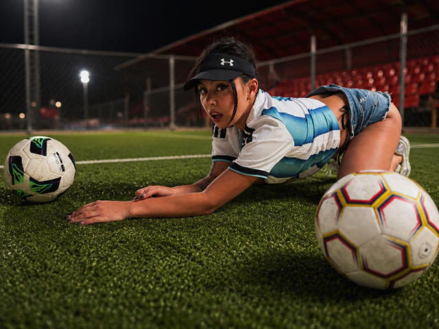 NenaBlack, hockend auf dem Gras, trägt eine schwarze Kappe, ein weißes und blaues Fußballtrikot und Jeansshorts, und hält zwei Fußballbälle, mit einem entschlossenen Gesichtsausdruck..