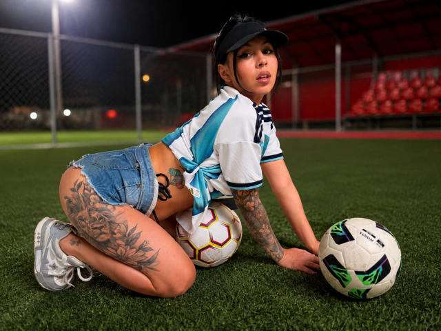 NenaBlack auf dem Rasen, kniend, trägt ein hochgebundenes Fußballtrikot, Jeansshorts, weiße Turnschuhe, eine schwarze Kappe, hat Tätowierungen am Oberschenkel und hält zwei Fußballbälle. Im Hintergrund ein Stadion bei Nacht, der Hintern ist sichtbar, es gibt Tätowierungen und sie hat einen sportlichen Look..