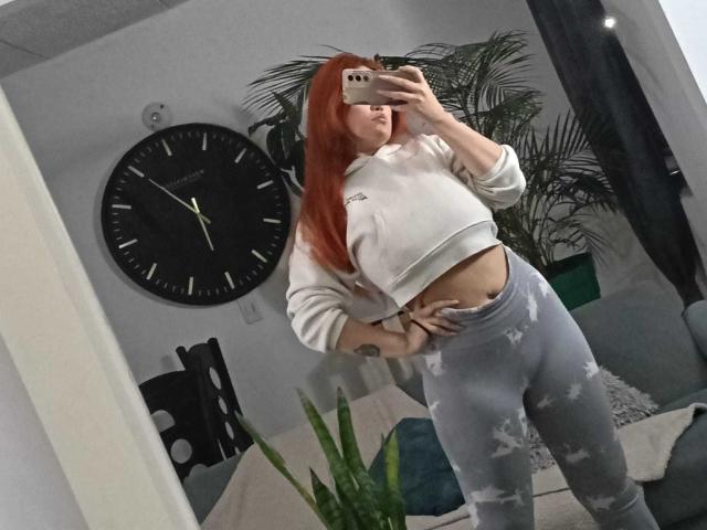 AmberLaurent, avec des cheveux roux, portant un sweat à capuche blanc et des leggings gris déchirés, montrant ses fesses, prend un selfie devant un miroir dans une pièce avec une horloge noire et une plante verte..
