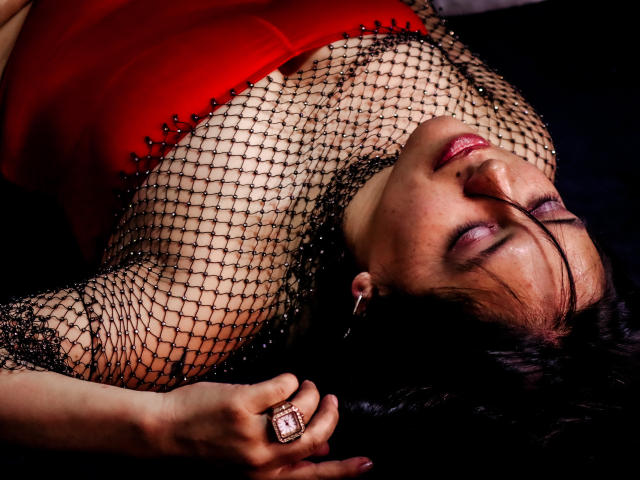 ZaharaViscon, con sus pechos apenas cubiertos por una camiseta de malla negra, un vestido rojo y un anillo, acostada con los ojos cerrados, cabello oscuro y una expresión sensual..