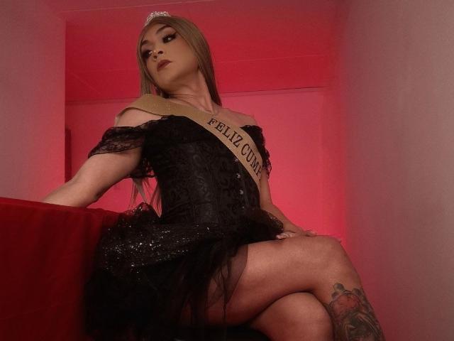Karol, elegant mit langen blonden Haaren, in einem schwarzen Spitzenkleid und einer "FELIZ CUM"-Schärpe, sitzt auf einem roten Sofa, die Beine gekreuzt, mit einem sichtbaren Tattoo am rechten Oberschenkel, unter rotem Licht..