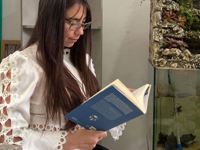 FreyaraVenwo, met lang bruin haar, een bril en een witte kanten jurk, leest een blauw boek. Ze heeft kleine borsten en een schattige achterkant, en staat in de buurt van een aquarium..
