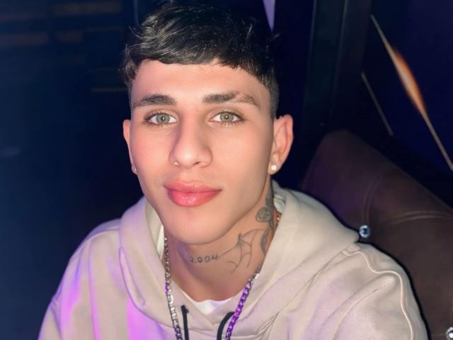 DanntheStiff with short dark hair, green eyes, and a white hoodie, has a nose ring and tattoos on his neck, sitting in a dark, lit-up area.
