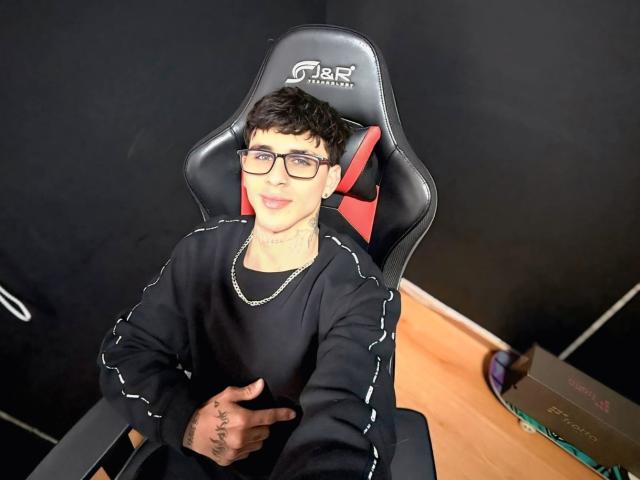 DanntheStiff with glasses, black sweatshirt, and necklace, sitting in a black gaming chair with red accents, smiling, tattoo on right hand, wooden floor, box in background.