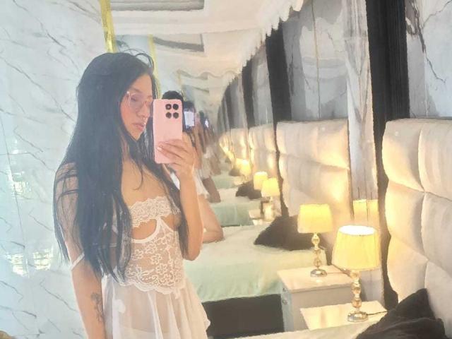 SophieDelaney with long black hair, wearing a white lace babydoll, showing her tits in a mirror selfie, holding a pink phone, in a luxurious bedroom with lamps.
