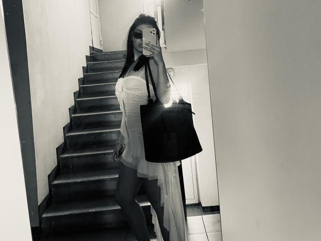 Selfie in bianco e nero di AlexXas con lunghi capelli scuri, che indossa un abito bianco trasparente, tiene una borsa nera, è in piedi su delle scale scure, mostrando il sedere e le gambe scoperte..