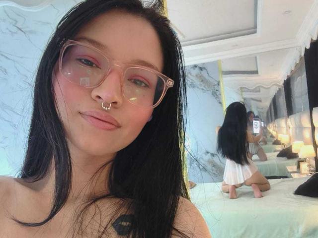 SophieDelaney with long black hair, pink glasses, nose ring, and pink eyelids, pouting with her tongue slightly out, wearing a white dress, kneeling on a bed.