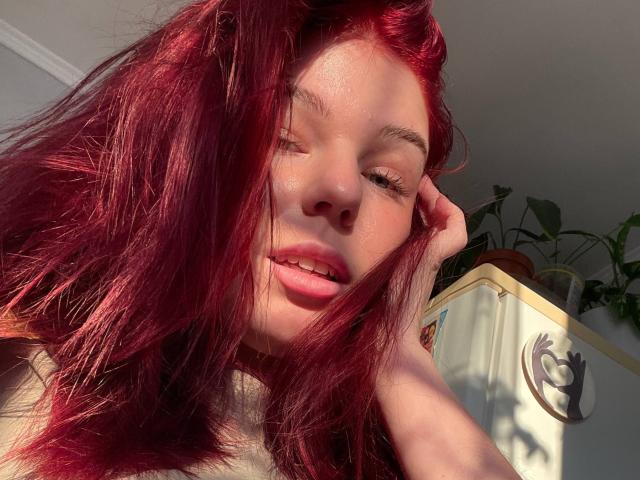 AlexXas ha i capelli rossi, la pelle illuminata dal sole, e si sta toccando la testa con un'espressione rilassata, con la bocca leggermente aperta. Le sue labbra rosa e il trucco leggero sono visibili..
