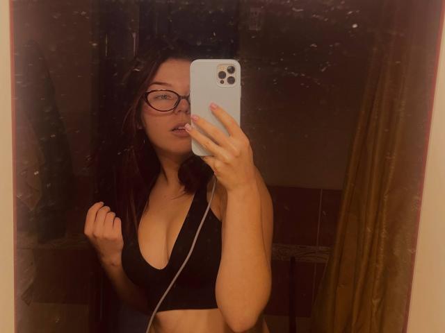 AlexXas with glasses, black sports bra, showing tits, holding phone, mirror selfie, dark hair, pink lip, slightly open mouth, reflection, dark room, yellow curtain, selfie, iPhone.