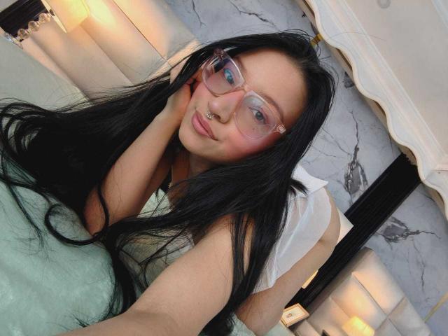 SophieDelaney with long black hair, pink glasses, nose ring, and white top, lying on a bed, green pillow, smiling, touching her face, soft lighting, bedroom.