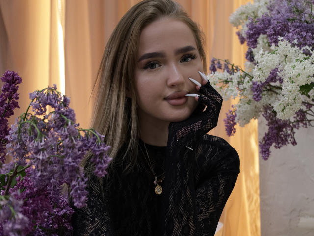AnfisaRalphs with blonde hair, wearing a black lace top, long white nails, and a gold necklace, holding purple and white lilacs, smiling softly.