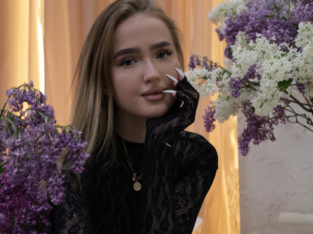 AnfisaRalphs, con cabello castaño claro, vestida con encaje negro, tocándose la cara con uñas largas y blancas, rodeada de flores moradas y blancas, mirando a la cámara con una sonrisa sutil..