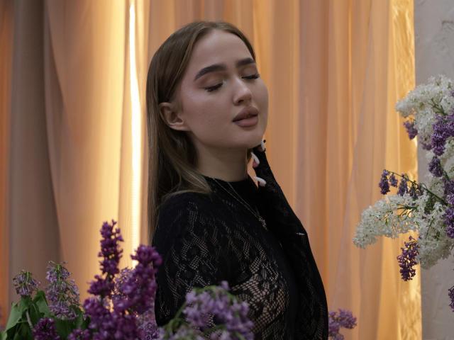 AnfisaRalphs with her eyes closed, wearing a black lace top, standing among purple and white flowers, her ass slightly visible, brown hair, soft light, serene expression.