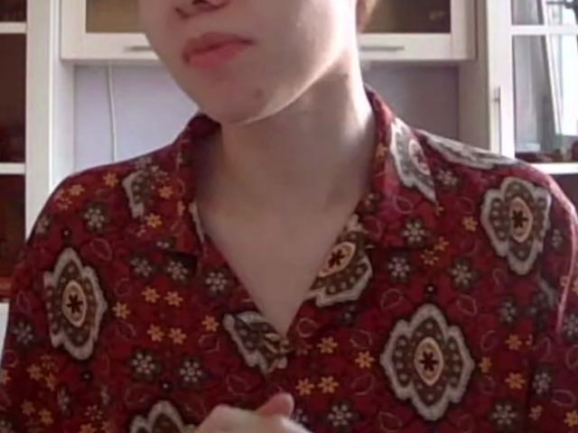 El rostro de Carmelita Bemiller es parcialmente visible, lleva una camisa roja con estampado floral en blanco y amarillo, el cuello está ligeramente abierto, tiene la piel clara y sostiene algo cerca del pecho..