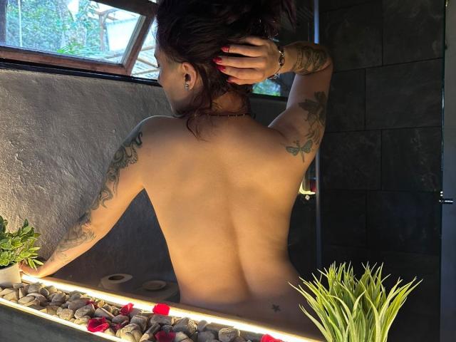 LiaMiss, topless, facing away, showing tattoos on arms, hands in dark hair, in a sunlit stone bathtub with plants and pebbles. Her back, ass, and tattoos are visible.