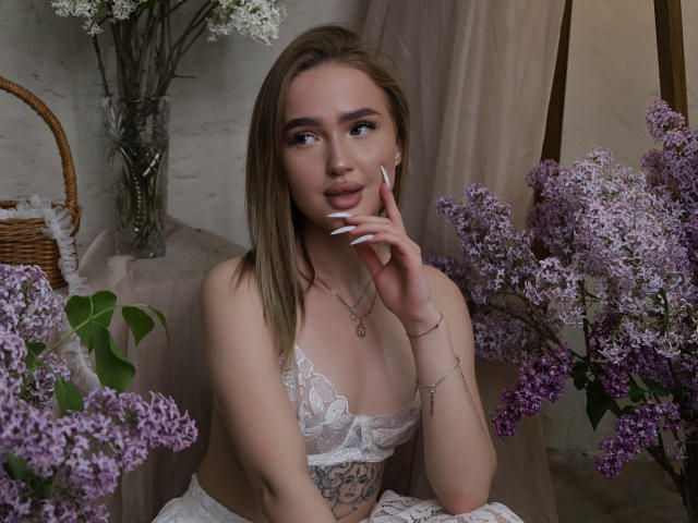 AnfisaRalphs con flores lila, blusa blanca de encaje, uñas largas, tatuajes en el lateral, mano en la mejilla, mirando hacia arriba, cabello rubio, piel pálida, maquillaje sutil, sonrisa suave, cesta de mimbre en el fondo..