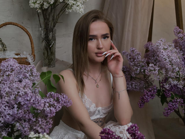 AnfisaRalphs, con lilas moradas, vistiendo lencería blanca de encaje, tiene senos de tamaño medio, piel clara, cabello castaño y un tatuaje en la muñeca derecha, tocándose los labios con un dedo..