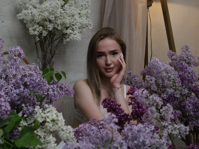 AnfisaRalphs, avec des lys pourpres et blancs, tenant un téléphone à l'oreille et souriant. Elle a des seins, des cheveux bruns clairs et un joli visage, et elle porte un haut blanc..