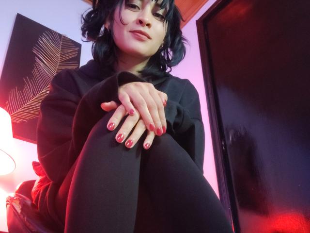 IssisDiosa, com cabelos escuros e ondulados, moletom preto e calças legging pretas, sentada com as mãos nos joelhos, esmalte vermelho, sorriso discreto, olhando para baixo, em iluminação rosa e vermelha suave..