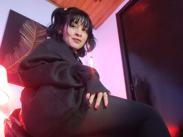 IssisDiosa with black hair, pigtails, black hoodie, and black leggings, sitting with one hand on her thigh, red nails, smiling, in a dimly lit room.