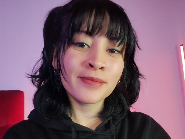 IssisDiosa mit schwarzen Haaren, Pony und einem leichten Lächeln, trägt eine schwarze Kapuzenjacke, hat eine Nasenring und blickt mit rosafarbenem Licht auf die Kamera..