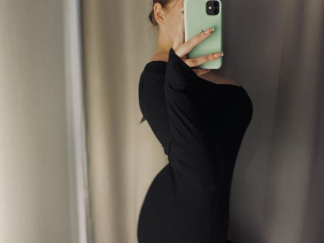 MissKuromi in a black long-sleeve dress, holding a green phone, covering her face, showing her curvy body and prominent tits in a mirror selfie.