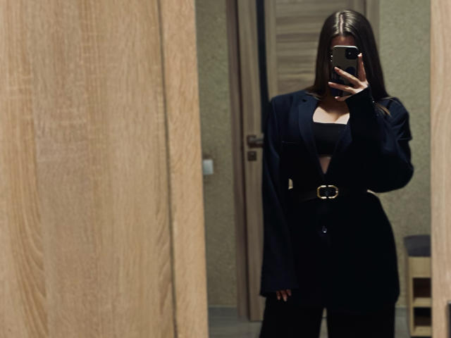 Mirror selfie of MissKuromi with long brown hair, black outfit, and belt, holding phone, covering face, showing small tits, and a small ass, in a wood-paneled room.
