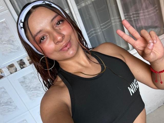 Saarah Jones met nat bruin haar, een zwarte sportbeha, oorringen en een rode armband, die een vrede-teken laat zien, waarbij haar kleine borsten en een lichte grijns zichtbaar zijn..