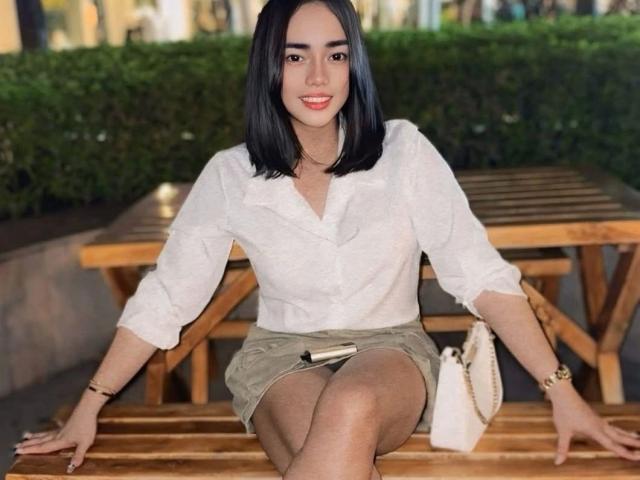 KylieSELFSUCKER-hot with black bob, white blouse, beige skirt, and white purse, sitting on wooden bench, smiling, legs crossed, hands on bench, wrist bracelet, wooden table behind.