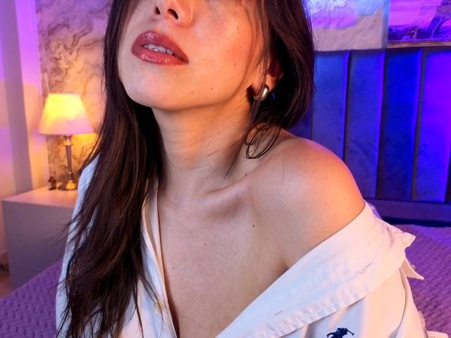 MiaReed with glossy red lips, slightly parted, wearing a white off-shoulder shirt, brown hair, blue light, and a gold earring, her tits subtly visible.