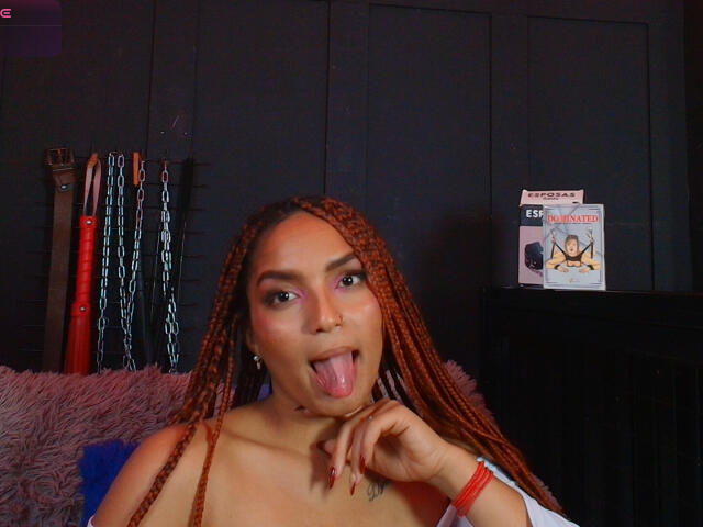 BrianaPratt with long orange braids, sticking out her tongue, has tan skin, red nails, and a red bracelet, wearing a white top, showing her tits.