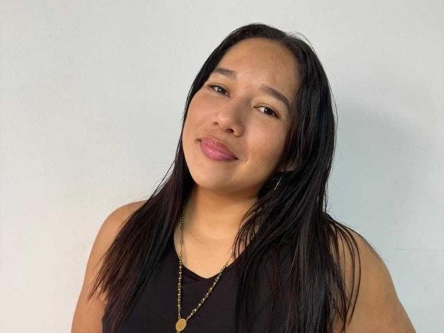 Chanttal Williams, avec ses cheveux noirs lisses, sa peau de couleur moyenne, ses lèvres roses, portant un haut noir sans manches et un collier en or, sourit légèrement. Ses seins sont de taille moyenne, et elle a l'air adorable..