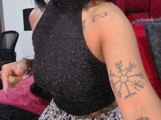 Amelie Blake, con tatuaggi sul braccio e sulla spalla, indossa una canottiera nera scintillante, tiene in mano un'unghia rosa, mostrando un tatuaggio a forma di fiocco di neve e un simbolo dell'infinito..
