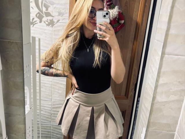 SabrinaDesire with long blonde hair, glasses, black sleeveless top, tattooed arm, and beige pleated skirt, taking a mirror selfie, showing her tits and ass.
