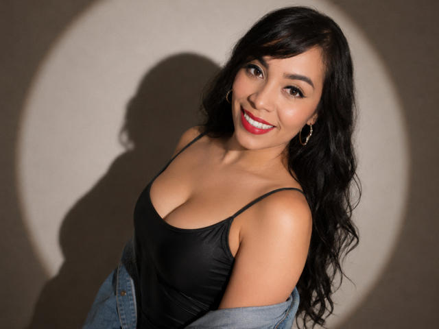 FetishLoverAna con canotta nera, che mostra il seno, sorridente, rossetto rosso, lunghi capelli neri, jeans portati sulle spalle, ombra sul muro, orecchini, carina, sexy, asiatica, sicura di sé..