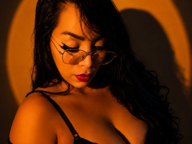 FetishLoverAna, avec ses cheveux noirs et bouclés, ses lunettes, son rouge à lèvres rouge et son soutien-gorge noir, regarde vers le bas, les yeux fermés, dans une lumière orange chaude qui met en valeur ses seins volumineux et ronds..