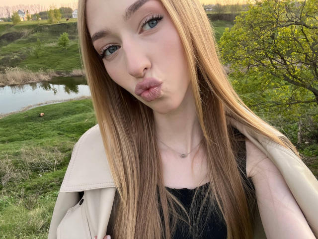 DionHolster has long blonde hair, green eyes, and pouts her lips. She's wearing a beige jacket over a black top. Her nose is pierced. Her ass is not visible.