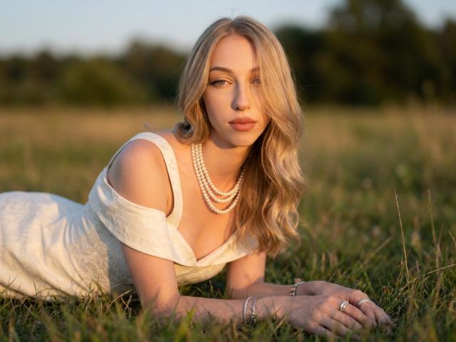 DionHolster, blonde woman, laying in grass, white off-shoulder dress, pearl necklace, wavy hair, soft gaze, sunlight, ring on finger, bracelet, serene expression, attractive, subtle makeup.