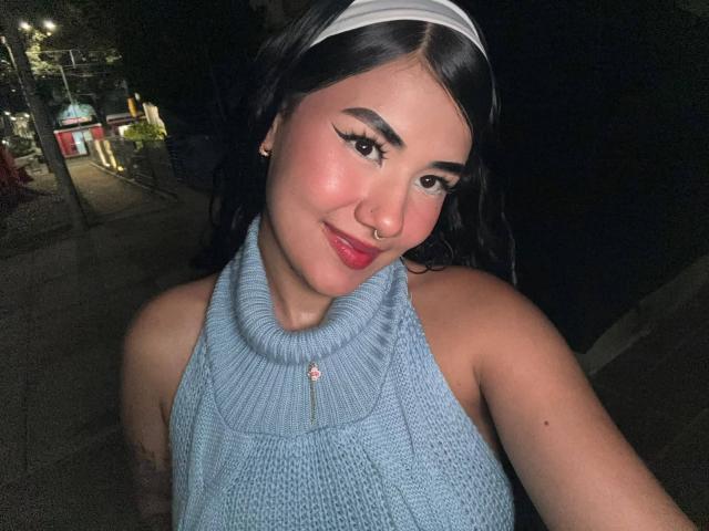 CuteAurora con cabello oscuro, diadema blanca, top de punto azul tipo halter, piercing en la nariz y lápiz labial rojo, sonriendo a la cámara, de noche, con un tatuaje visible en el brazo..